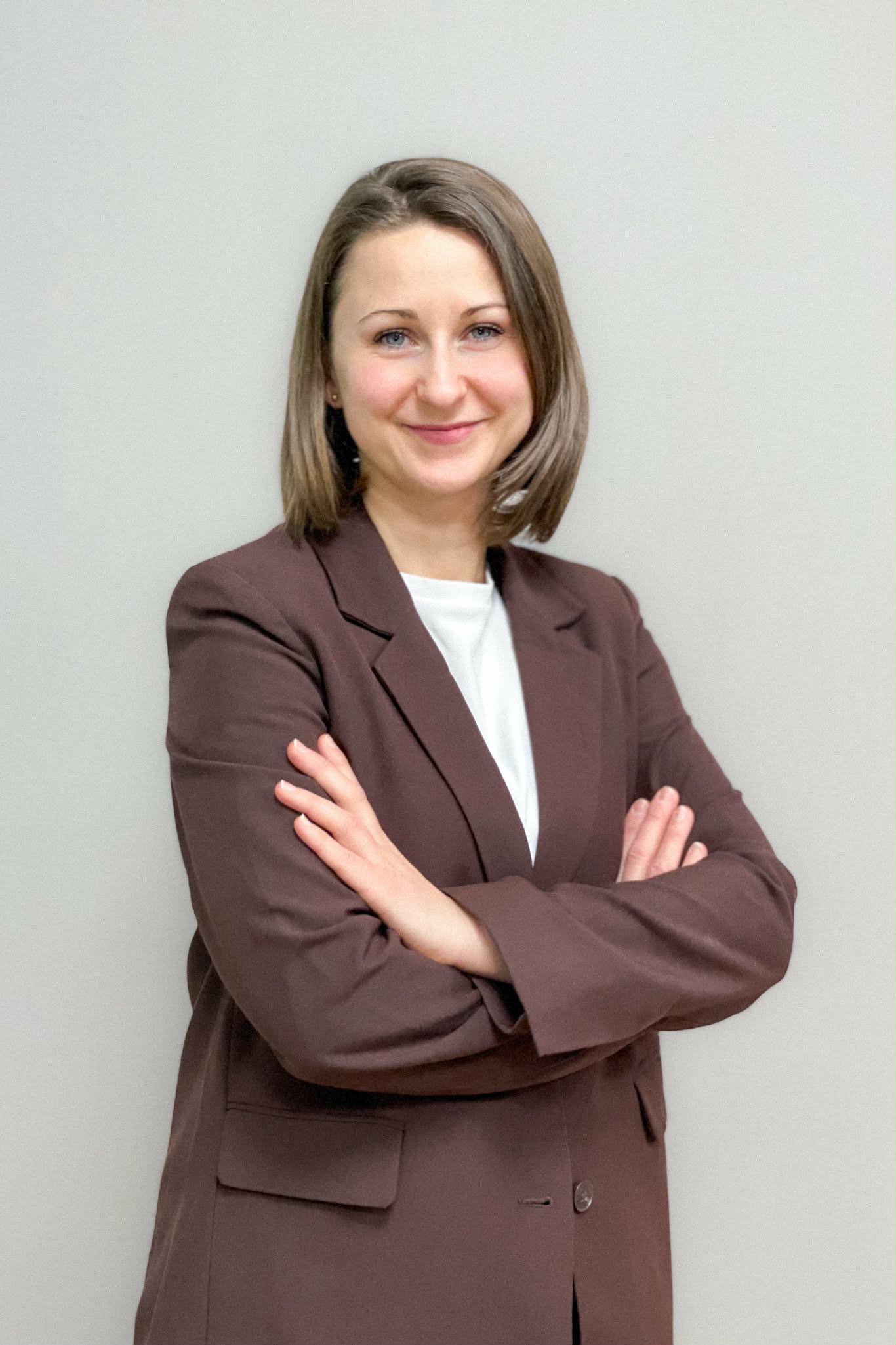 Uśmiechnięta kobieta z krótkimi włosami stoi z założonymi rękami, ubrana w brązową marynarkę i jasną bluzkę, na tle jasnej ściany.