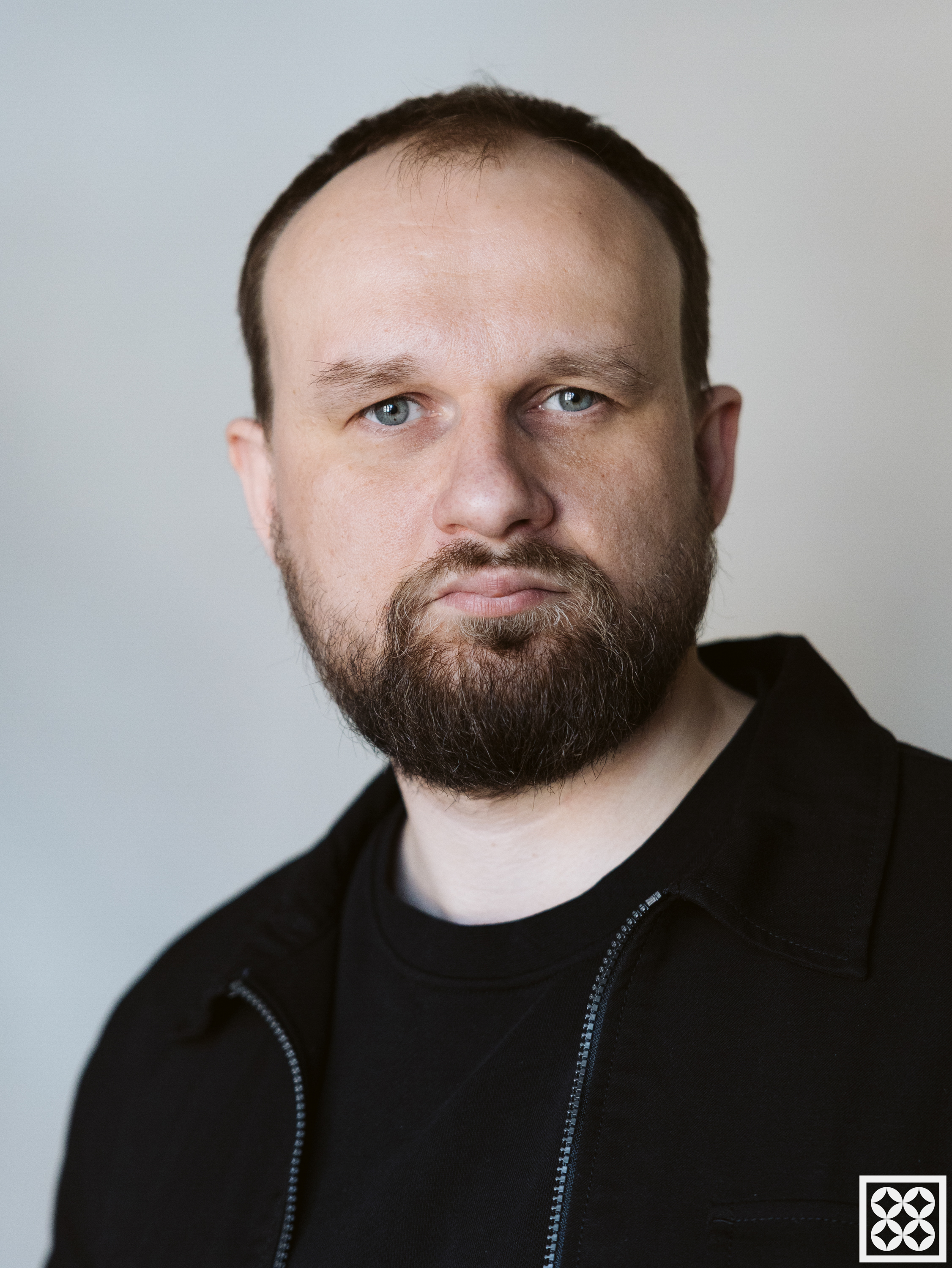 Portret mężczyzny z brodą na jasnym tle, w czarnej bluzie.