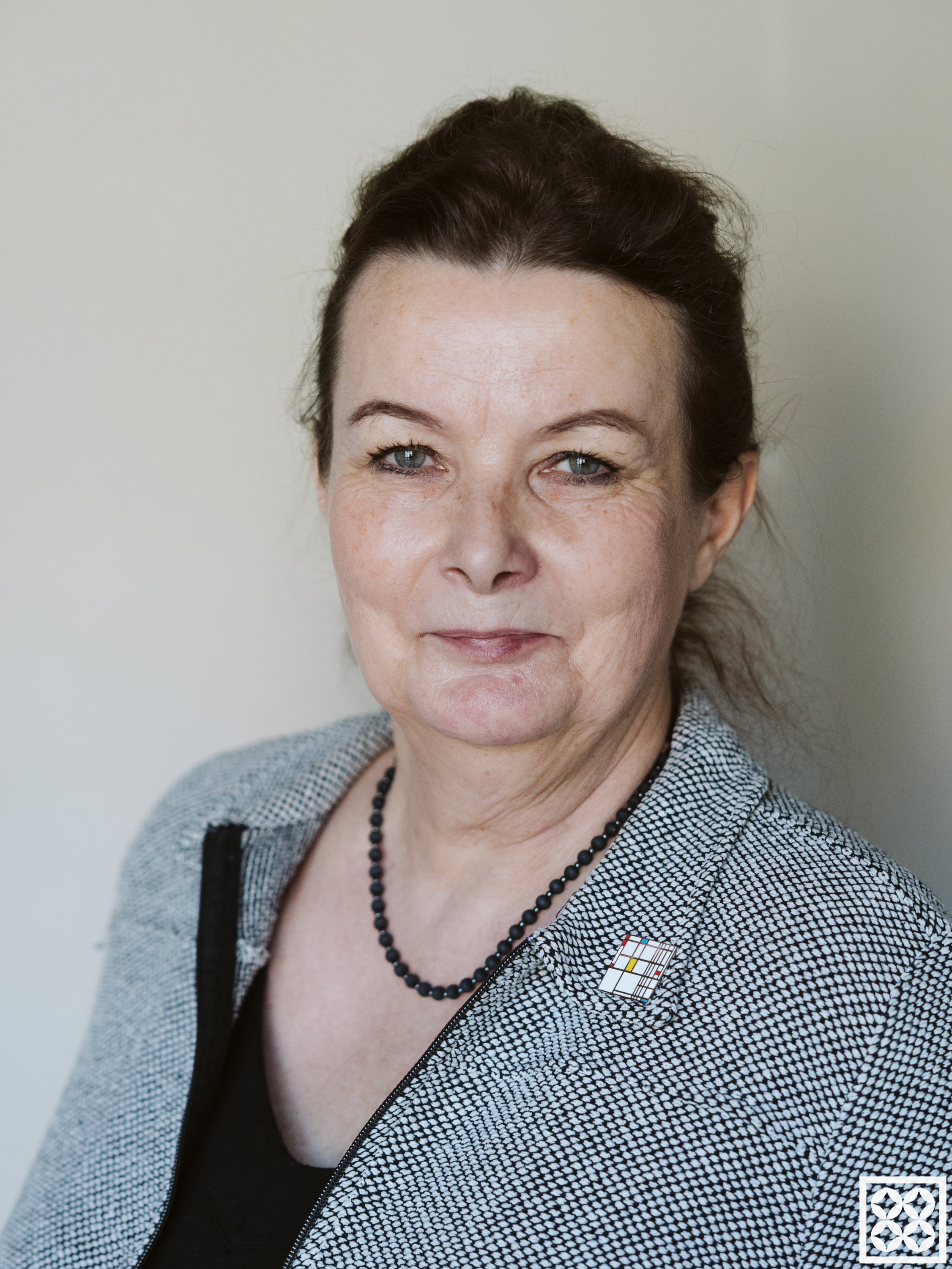 Portret kobiety w średnim wieku na jasnym tle, z lekkim uśmiechem, w szarej marynarce i czarnym naszyjniku.