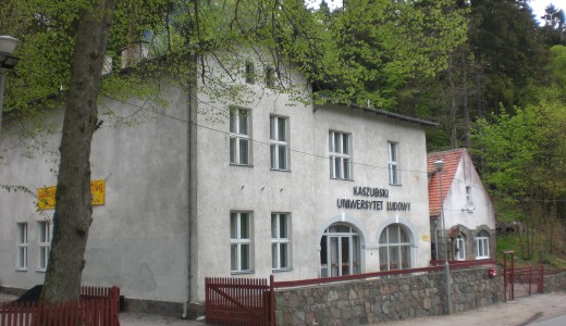asny, dwupiętrowy budynek z napisem „Kaszubski Uniwersytet Ludowy”, położony przy ulicy wśród gęstych, zielonych drzew. Przed budynkiem znajduje się niski kamienny mur i czerwone ogrodzenie, a całość otacza leśne otoczenie.