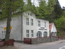 asny, dwupiętrowy budynek z napisem „Kaszubski Uniwersytet Ludowy”, położony przy ulicy wśród gęstych, zielonych drzew. Przed budynkiem znajduje się niski kamienny mur i czerwone ogrodzenie, a całość otacza leśne otoczenie.